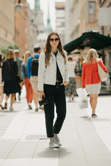 A gorgeous young female blogger in a white jeans jacket, black jeans, grey sneakers, and sunglasses holds the tripod with smartphone poses in the center of busy ancient European street