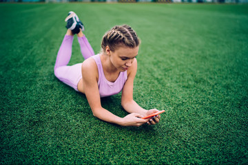 Woman on grass of field using smartphone.From above serious sportive woman lying on green grass with crossed legs and browsing mobile phone preparing for training on athletic field