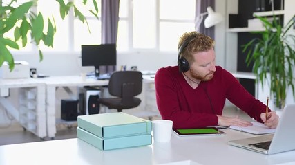 Young handsome innovative caucasian freelancer sitting in his office with headphones on, watching tutorial on tablet and writing in notebook result of his research.