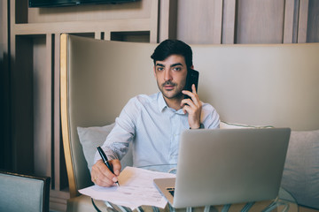Portrait of latin male director in elegant formal wear writing during mobile phone conversation using modern technology, prosperous businessman making cellular call writing information about project.