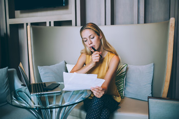 Concentrated blonde woman in smart casual wear pondering while checking planning strategy for project holding paper, millennial female manager checking data using modern technologies for research. © BullRun