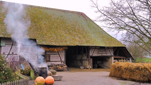 Granja r&uacute;stica de fachada blanca y gran tejado verde cocinando en el exterior caldero humeante de calabaza y verdura