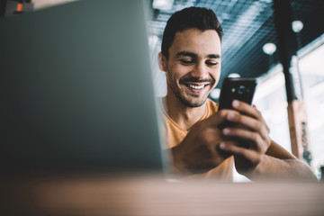  Millennial male laughing watching funny video on smartphone spending free time in cafe interior,successful man blogger satisfied with online earning getting income on banking account on cellular.