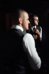 The groom looks in the mirror. Portrait of a stunning groom dressing up for the wedding and getting ready in his room.