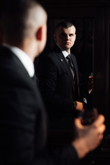 The groom looks in the mirror. Portrait of a stunning groom dressing up for the wedding and getting ready in his room.