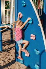 Fototapeta na wymiar Little preschool girl climbing rock wall at playground outside on summer day. Happy childhood lifestyle concept. Seasonal outdoors activity for kids. Strong girl female power.