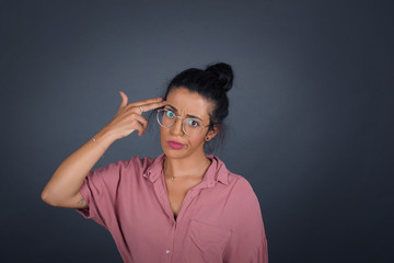 Unhappy blonde young female imitates gun shoot makes suicide gesture keeps two fingers on temples has puzzled expression dressed in casual clothes stands indoors.