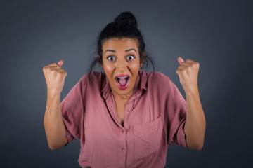 Waist up portrait of strong successful determined young female winner in casual t-shirt raising arms, clenching fists, exclaiming with joy and excitement. Victory, success and achievement concept.