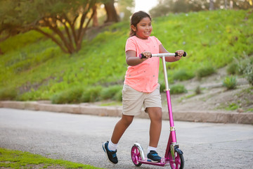 A little girl riding a pink scooter with confidence in a street lined with trees and grass.