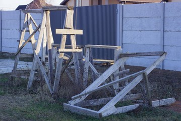 Fototapeta premium a pile of gray wooden construction goats stands on the green grass on the street at a concrete fence and an iron gate