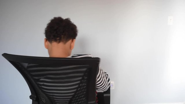 A young black african american pre teen showing different faces and turning around on his chairl. A cute playful boy, having fun, spinning his arm around. A young mixed male playing with by himself.