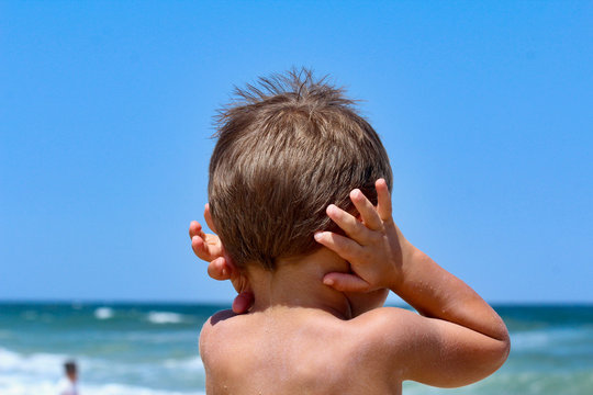 Young boy standing at the ocean with hands over ears during a quiet summer beach moment, capturing childhood calm, individuality, and a peaceful seaside atmosphere