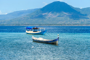 Two boats anchored to shallow waters of a small island near Maumere, Indonesia. Clear, turquoise coloured water displaying coral reef. There is a massive island in the back. Serenity and calmness.