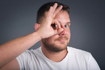 Fototapeta premium Gorgeous young blond man with happy face smiling doing ok sign with hand on eye looking through finger. Wearing casual clothes and standing.