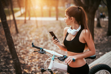 sports girl with headphones stands with phone and looks out into the distance near a bicycle at...