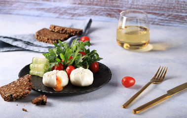 Breakfast with two poached eggs with flowing yolk, with arugula salad,cucumbers  and cherry tomatoes, on a black plate, with fresh grain bread with seeds and a glass of white dry wine.
