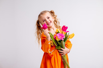 little blonde girl in an orange dress holds tulips and gerberas in her hands on a white background,...
