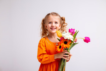 little blonde girl in an orange dress holds tulips and gerberas in her hands on a white background,...