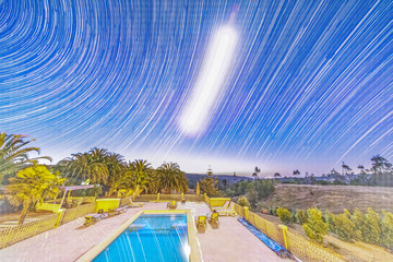 An amazing star trail nightscape with the Milky Way and its galactic core in Sagittarius below the shiny Moon light illuminating the Chilean countryside at a swimming pool on a rural Villa
