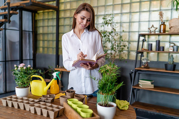 Landscape designer writing down research on plants in a notebook standing at a table with garden tools and garden plants in the winter garden