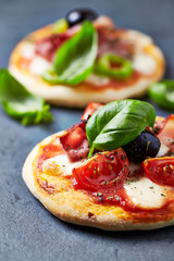 Mini pizzas with ham, mozzarella cheese, black olives and fresh basil on dark stone background. Close up.