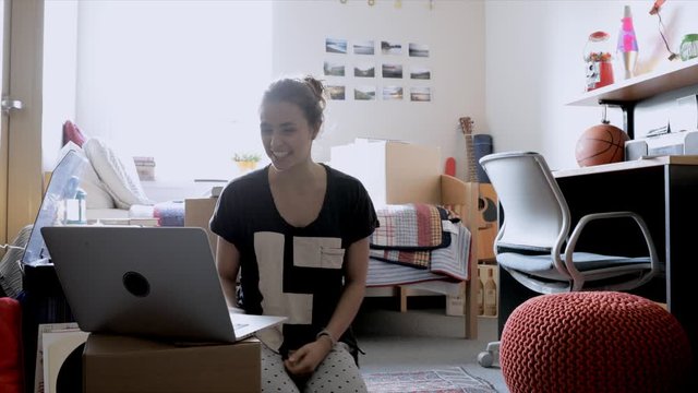 Female college student video chatting at laptop, moving into dorm room