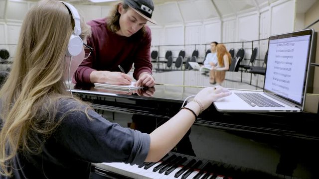 Music Students Writing Music On Laptop At Piano In Classroom