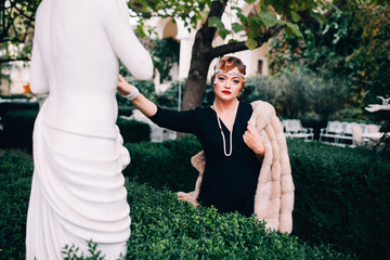 a gorgeous woman in a fur cape and a diadem on her head walks through her garden surrounded by...