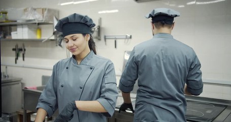 Cheerful young woman chef in working clothing and hat smiling and looking at camera while cooking at professional kitchen with male colleague standing behind. Concept of culinary and cuisine