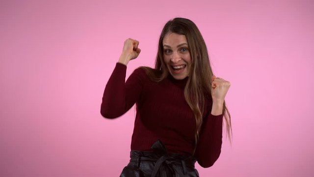 Happy smiling girl showing gesture of winner over studio pink background. Excited woman celebrating triumph and clenching her fists while looking at the camera. Dancing girl showing yeah gesture of