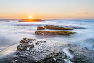 An amazing view of the sunset over the water in the Chilean coast. An idyllic beach scenery with the sunlight illuminating the green algae and rocks with orange tones and the sea in the background
