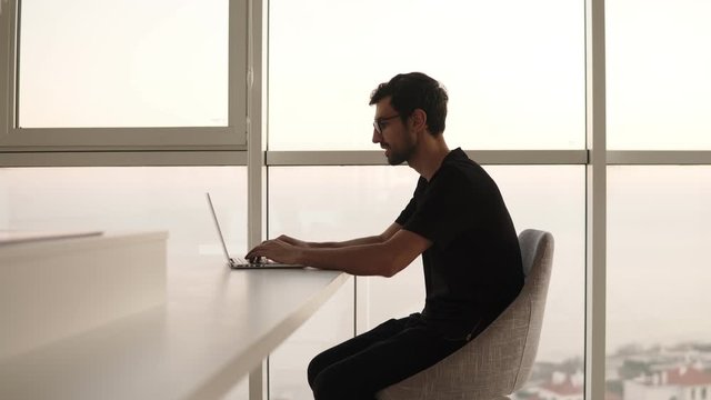 Side view of business man using laptop computer at remote workplace in slow motion. Freelancer working on computer at home. Focused man looking laptop at big house with panoramic windows