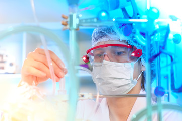 Young female scientist or tech in protective wear supervises chemical synthesis, checking reaction. Laboratory, glassware, futuristic turquiose light, research facility out of focus.