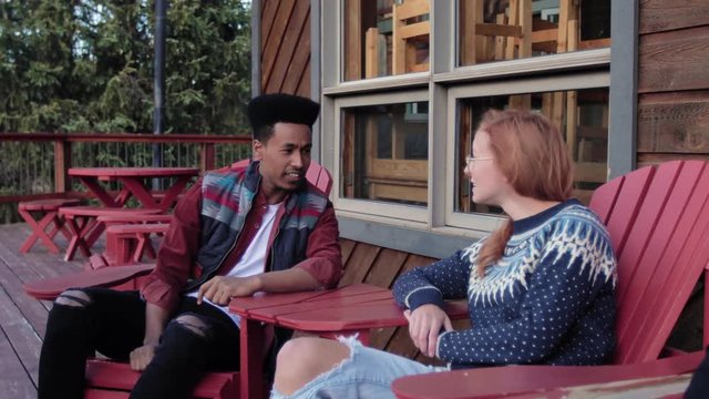 Young Couple Talking And Relaxing In Adirondack Chairs Outside Cabin