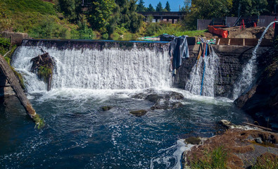 Thundering Upper Tumwater Falls pouring into the creek with a log and a bridge construction materials such as pipes and hoses plus a gray and red tarps surrounded by a fence with trees and grass