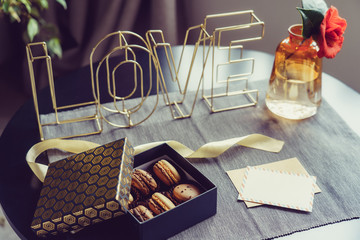 Festive box with chocolate macaroons cakes, rose flower in vase, postcard and wire word Love on gray napkin. Cozy gift idea for lover. Hygge Valentine's day concept. Romantic mood in noir nordic style