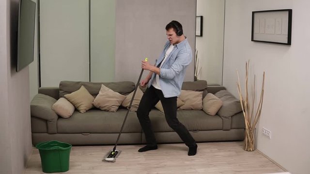 People, housework and housekeeping concept - happy man in headphones with mop cleaning floor and dancing at home. Using swab pipe as a guitar. Grey couch on the background