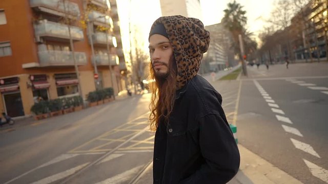 Portrait of long haired hipster walking on street in modern city in slow motion. He wears black jacket with leopard hoodie. Stylish man in sunset light