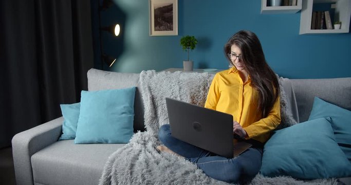 Pretty young lady with long dark hair in yellow blouse and blue jeans relaxing on grey couch at home and using laptop. Smiling woman in eyeglasses spending leisure time with computer