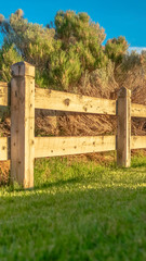 Vertical frame Brown wooden low fence on a lush green field against thick bushes and shrubs