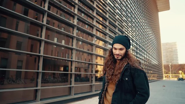 Portrait of long haired hipster walking on street in modern city in slow motion. He wears black jacket with leopard hoodie. Stylish man near business buildings
