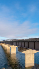 Obraz premium Vertical frame Bridge over a vast calm lake with snow covered shore during winter season