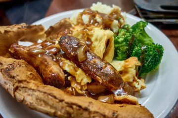 Close up of plate of food featuring toad in the hole a traditional British cuisine. Sausage and mash with broccoli, Yorkshire pudding, gravy and fried onions.     