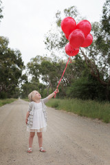 girl standing on dirt road holding red balloons