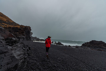 Obraz premium Girl with a red rain coat and a biene hat on Djupalonssandur black sand beach