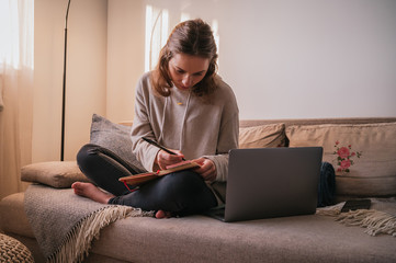 Home Office. Young business woman working at home with her computer and taking notes in her notebook, sitting in an armchair during sunset