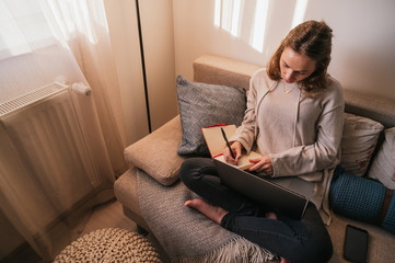 Obraz premium Home Office. Young business woman working at home with her computer and taking notes in her notebook, sitting in an armchair during sunset