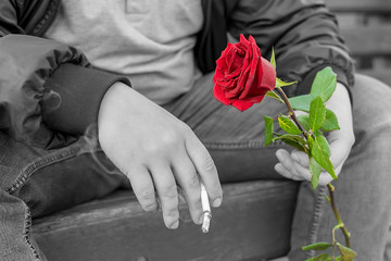 view of the hand of a man with a red rose in his hand, who is sitting with a smoldering cigarette on a bench in a city Park