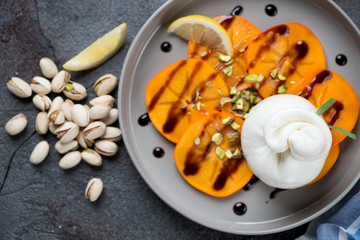 Salad with persimmon, burrata cheese and pistachios served on a grey plate, view from above, close-up
