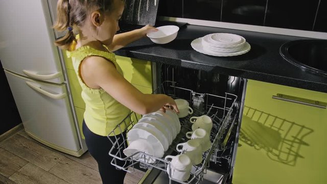 Smart girl learning to use dishwasher. Stylish modern Built In Kitchen Appliances in green black. Young mistress children study loading automatic electric dishwasher. Child is putting clean dishes.